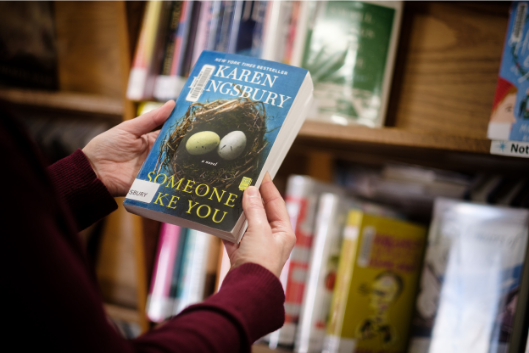 Person holding Karen Kingsbury's Someone Like You in front of library shelves