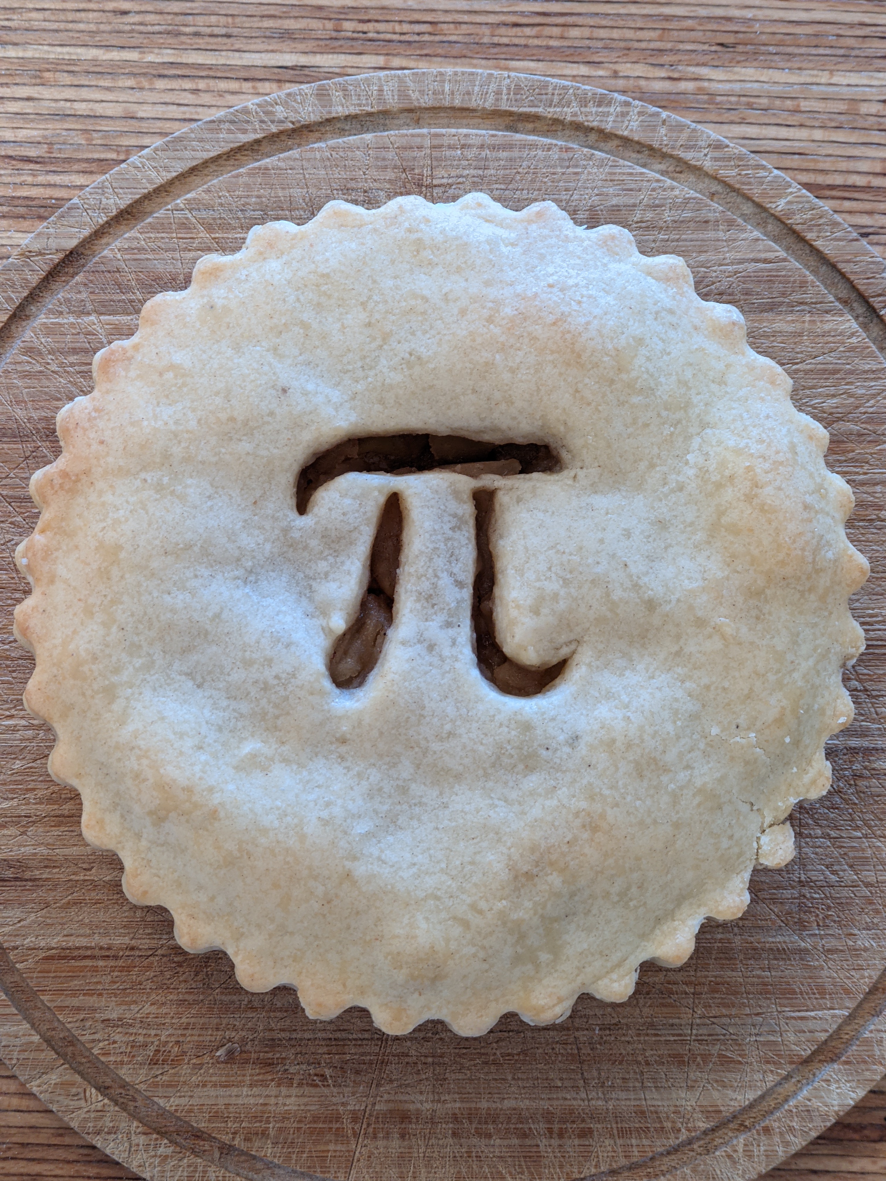 A photo of a pie on a wooden plate with the symbol for Pi in the center of the crust.