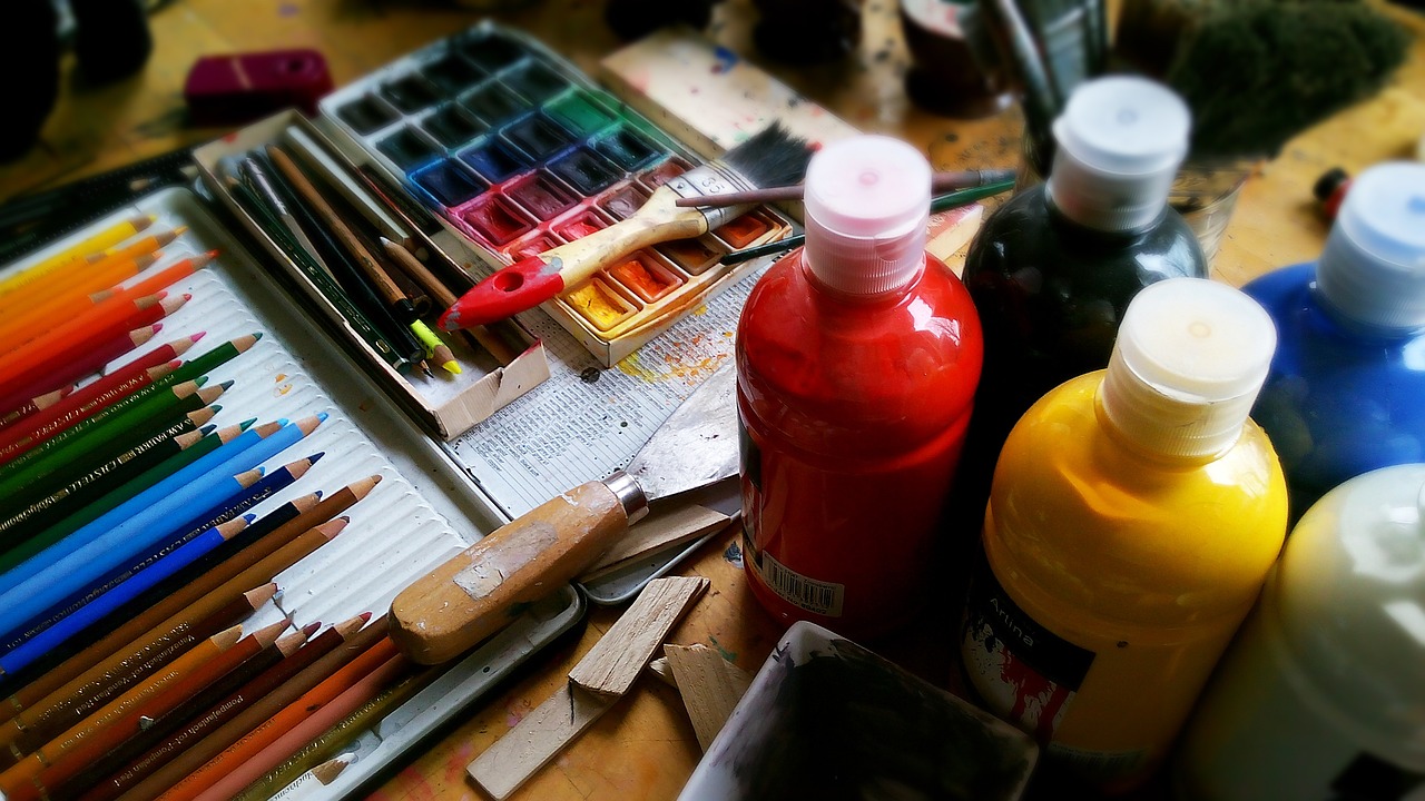 An image of art supplies including bottles of paint, watercolor paints, and colored pencils. 