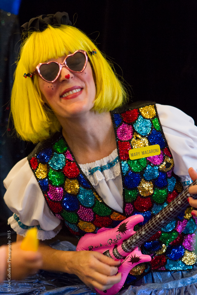 Mary Macaroni, woman in bright yellow wig playing inflatable toy guitar