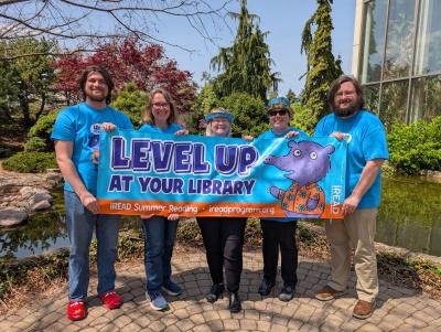 Librarians in matching Summer Reading shirts hold a Level Up at Your Library banner outside the QC Botanical Center
