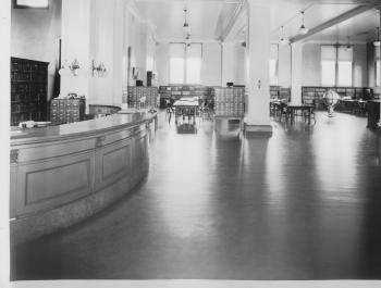Black and white photo of desks, card catalogs, and shelves of the early library interior