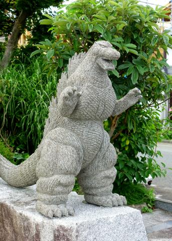 A grey stone statue of Godzilla stands in front of a tree with green leaves.