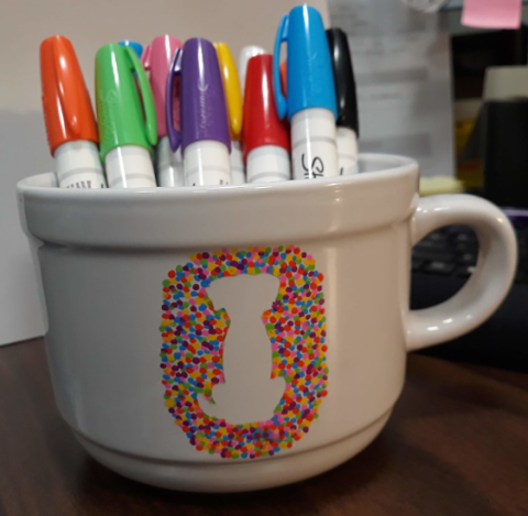A white mug with sharpie markers of different colors inside has been decorated with sharpie dots: together, the colorful dots form the outline of a dog.