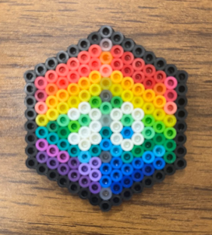 A rainbow D20 die made from perler beads sits on a faux wood table.
