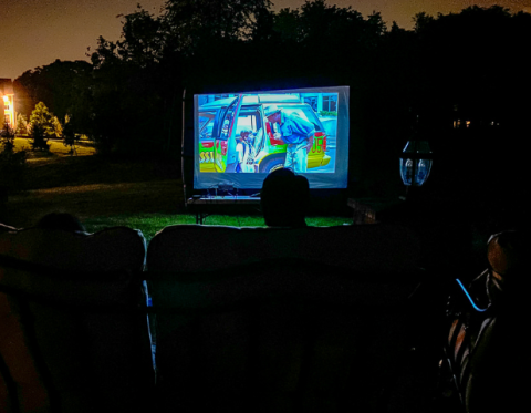 An inflatable movie screen appears surrounded by darkness with trees and a house in the background, and people in chairs in the foreground.