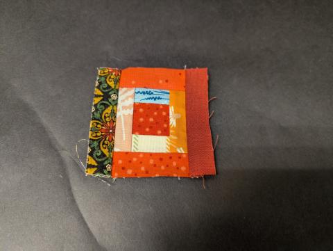 Very small quilt block made up of orange, peach, blue, and green strips in the shape of a log cabin.