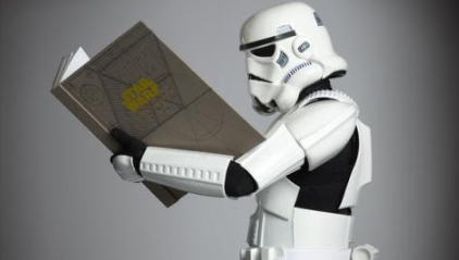 A stormtrooper from the Star Wars universe (in full white armor), holds a large beige book with "Star Wars" written in gold letters on the cover.