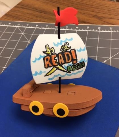 Toy foam pirate ship with blue background. Sail says 'Read! Argh!"