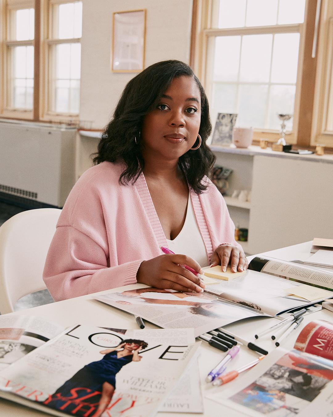 Photo of a young Black woman sitting at a desk covered in magazine spreads