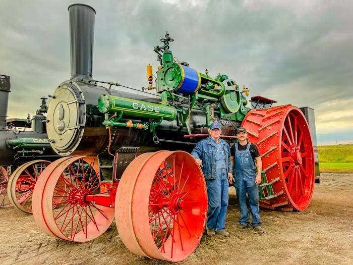 Men standing between front and back wheels of 150 Case - largest steam driven tractor