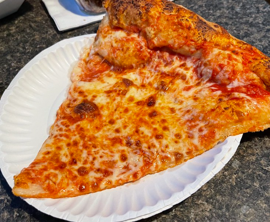A slice of cheese pizza on a white paper plate sits on a grey counter.