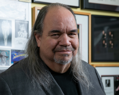 A headshot of a man in a grey suit, with a goatee and long grey-brown hair