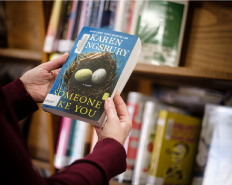 Person holding Karen Kingsbury's Someone Like You in front of library shelves
